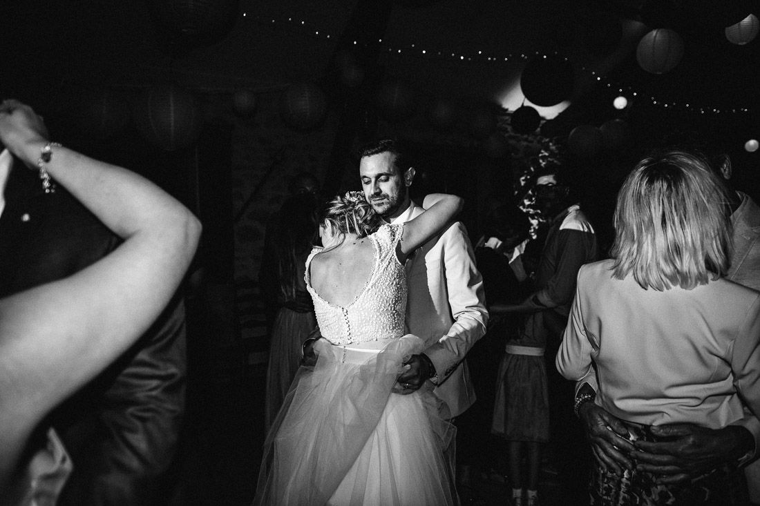 Black and white photo of bride and groom sharing emotional dance embrace surrounded by guests at wedding reception.