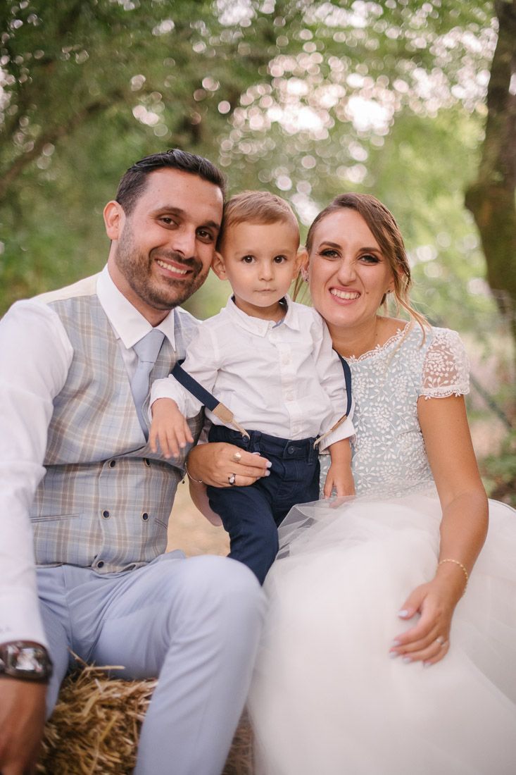 Smiling bride and groom pose with young son on hay bale at rustic outdoor wedding celebration.