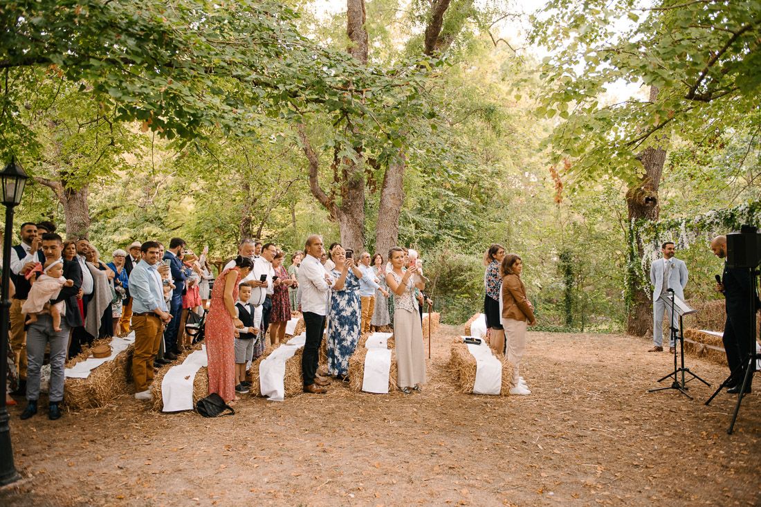 Outdoor woodland wedding ceremony with guests standing among hay bales beneath lush green trees.