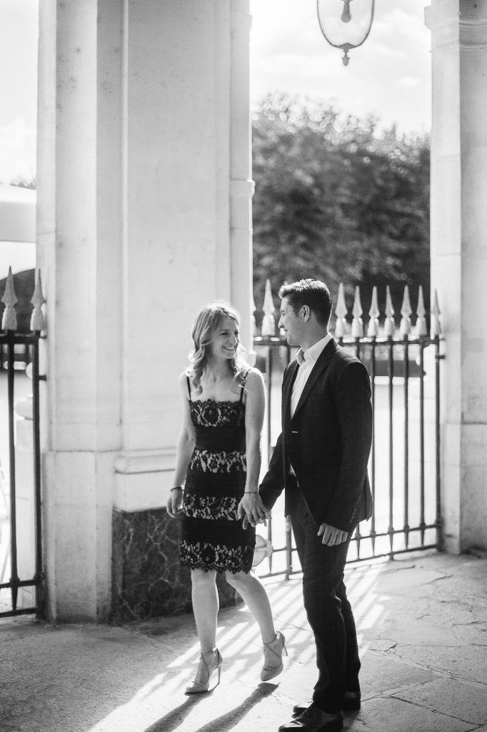 Couple holding hands walking through ornate columned archway in black and white engagement photo.