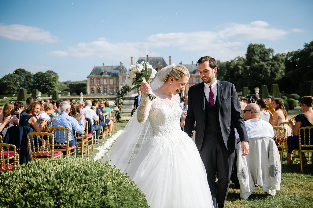 Smiling newlyweds walk hand in hand past guests after outdoor French chateau wedding ceremony.