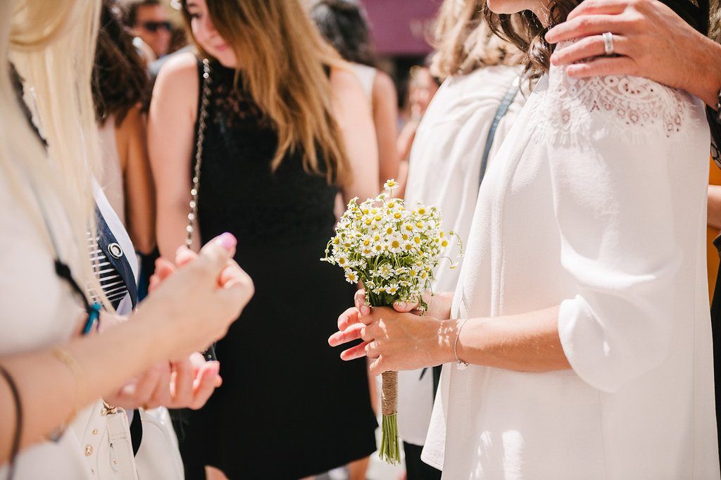 Bride holding a small daisy bouquet surrounded by guests at an outdoor wedding celebration.