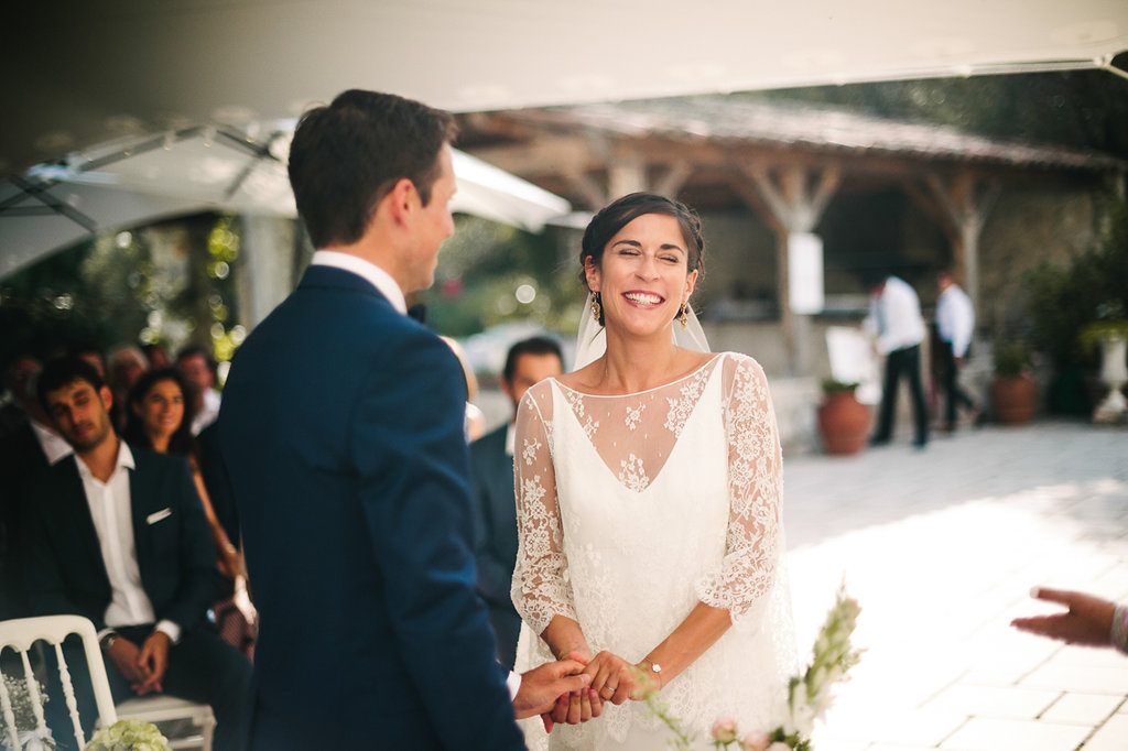Smiling bride holding grooms hands during outdoor wedding ceremony with seated guests applauding behind them.