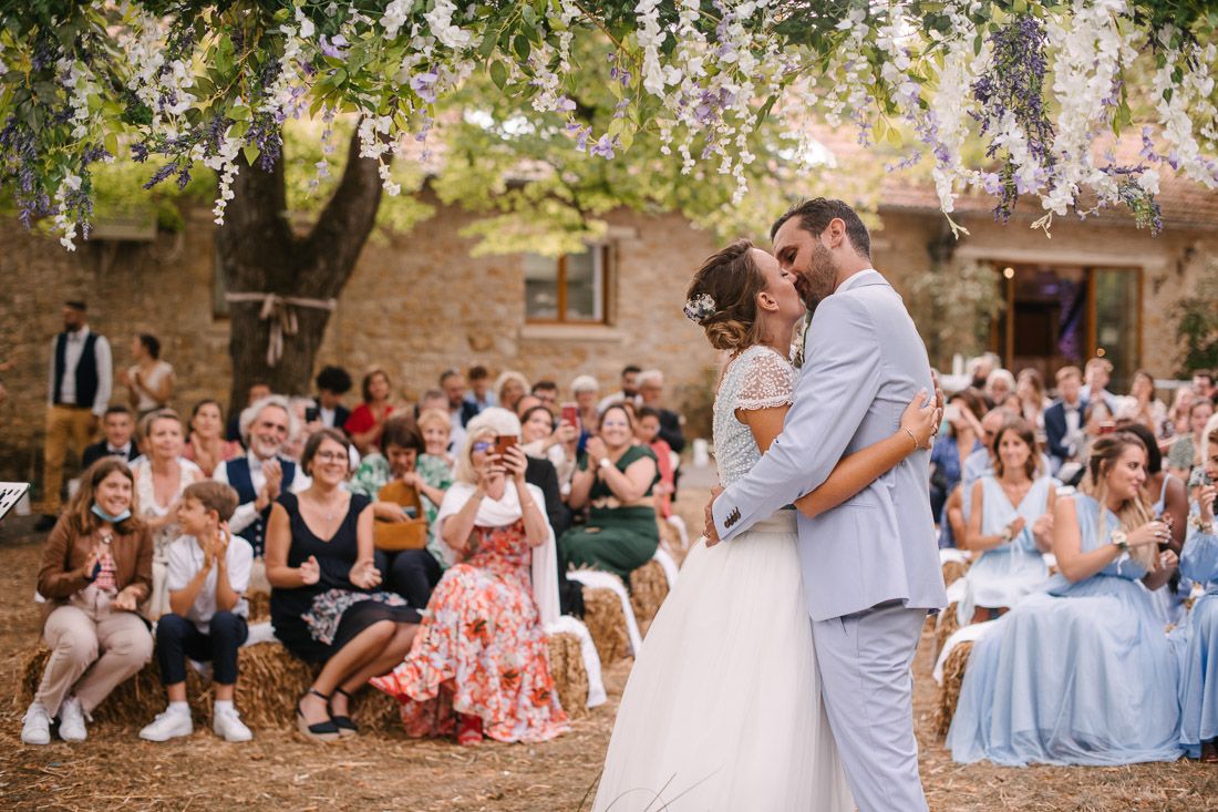 Romantic bride and groom embrace under flowering wisteria tree surrounded by wedding guests.