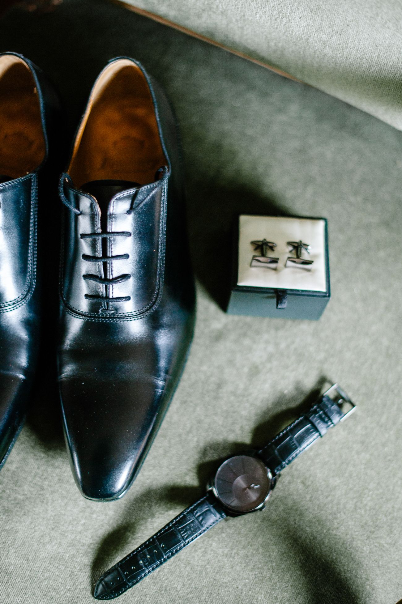 Black leather Oxford shoes with cufflinks in a box and a wristwatch arranged on a gray surface for a wedding.