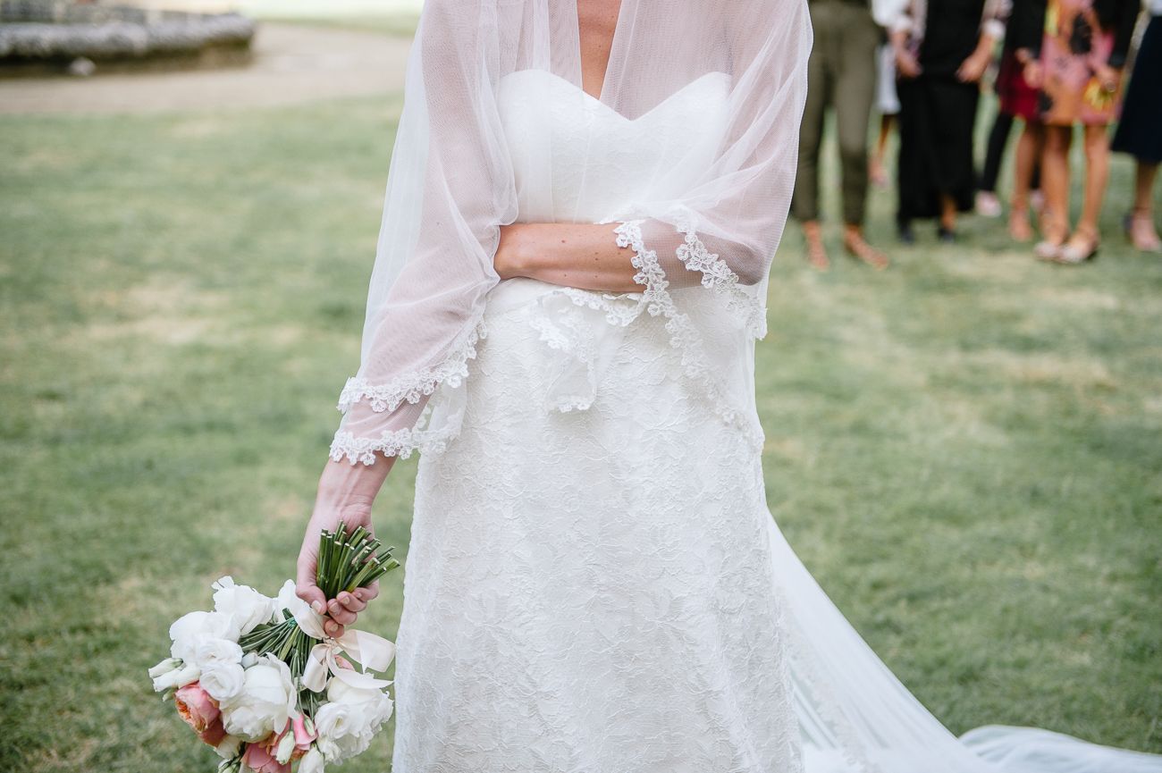 Close-up of bride in lace wedding gown with sheer wrap holding white and pink bouquet.