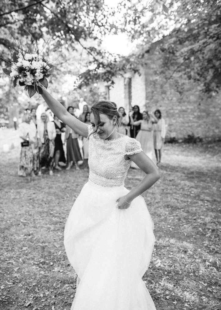 Black and white photo of bride tossing bouquet with guests watching in background at outdoor wedding.