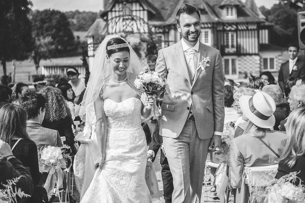 Radiant newlyweds walk hand in hand past cheering guests after outdoor wedding ceremony at elegant French estate.