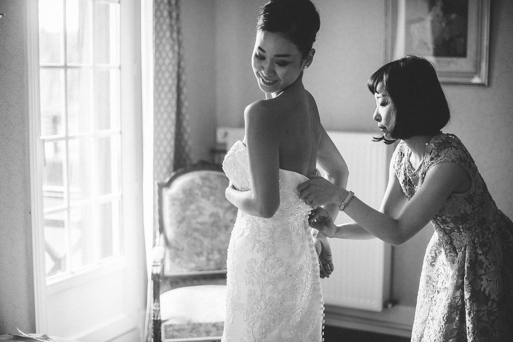Black and white photo of bride being helped into her lace wedding dress by a bridesmaid.