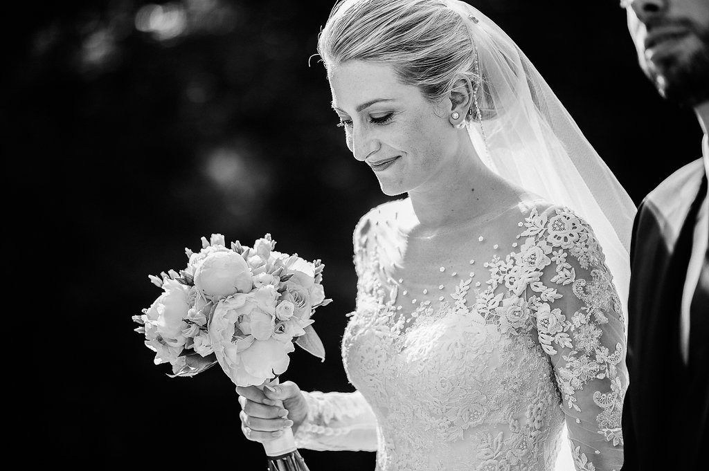 Bride in lace long-sleeve wedding dress holding bouquet, looking down in black and white portrait.