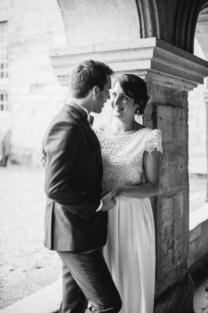 Bride and groom sharing a romantic glance leaning against a stone column, black and white.