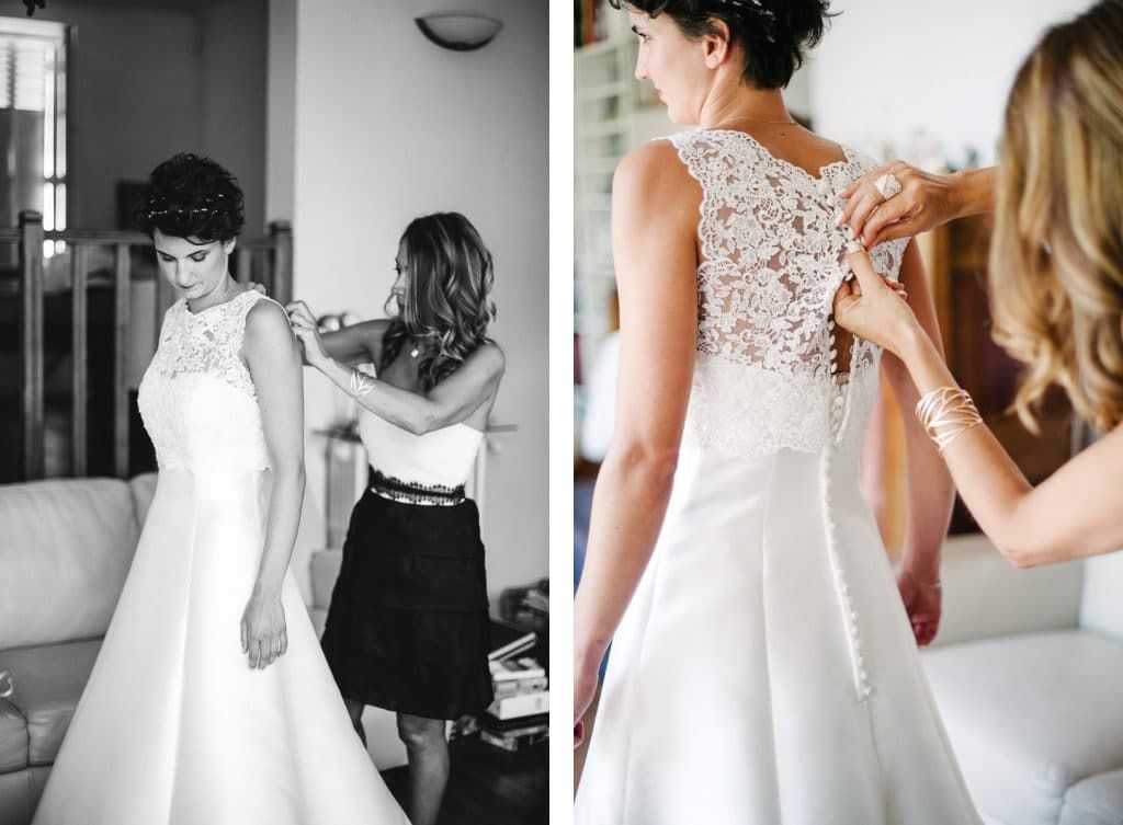 Bride getting dressed in white lace gown with bridesmaid helping button up the back before wedding ceremony.