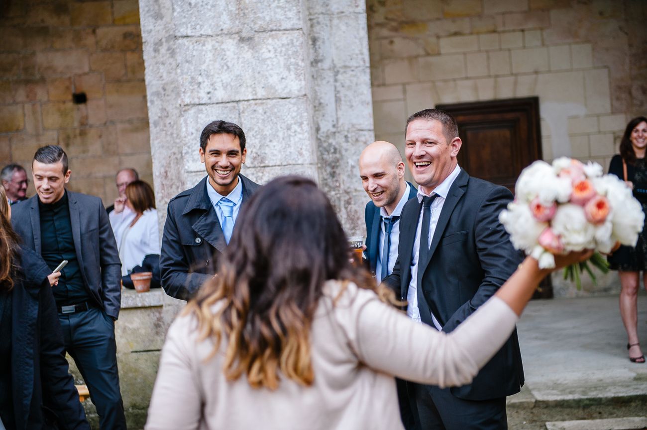 Wedding guests laugh and celebrate outside a stone venue as bride holds a pink bouquet.