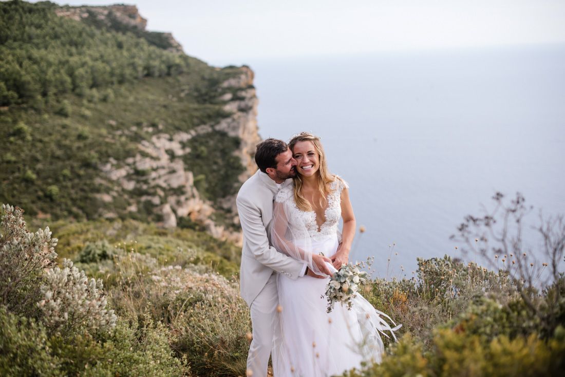Couple in wedding attire embracing on a scenic coastal cliff with ocean and rocky landscape in background.