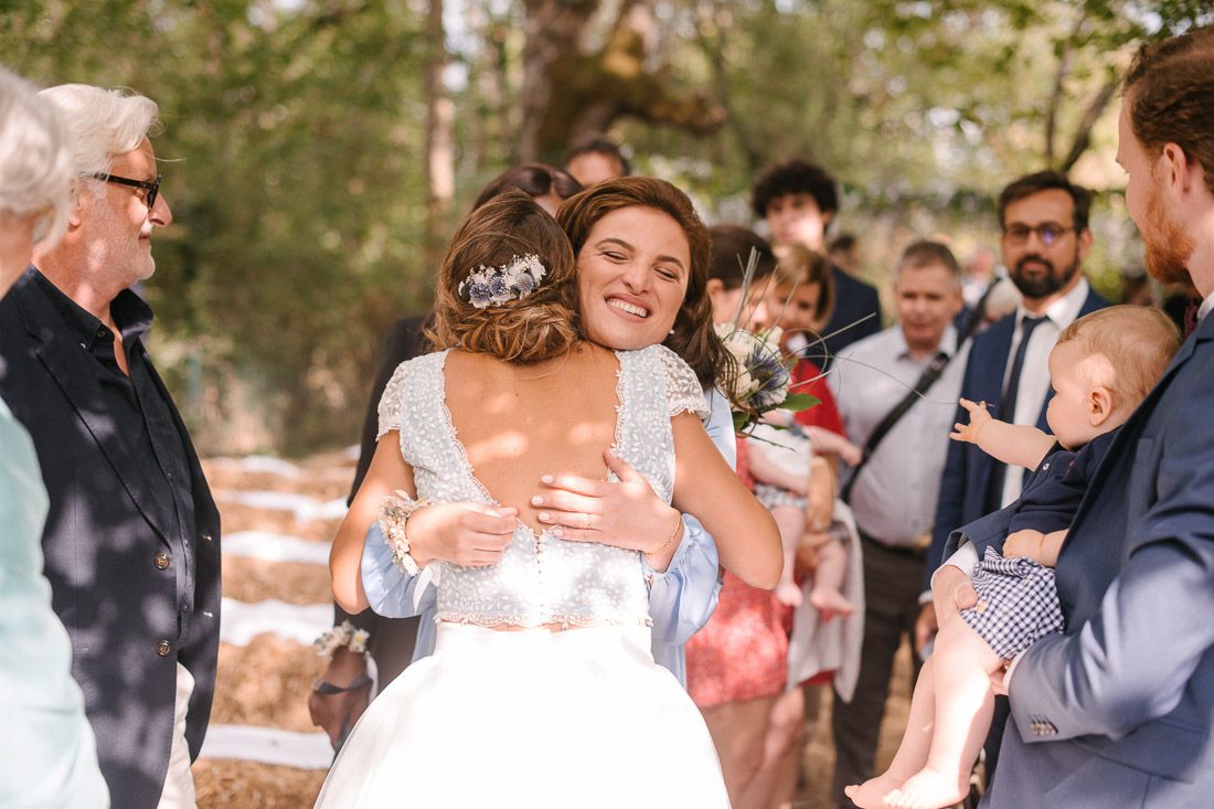 Smiling bride in lace wedding dress receives warm hug from guest after ceremony.