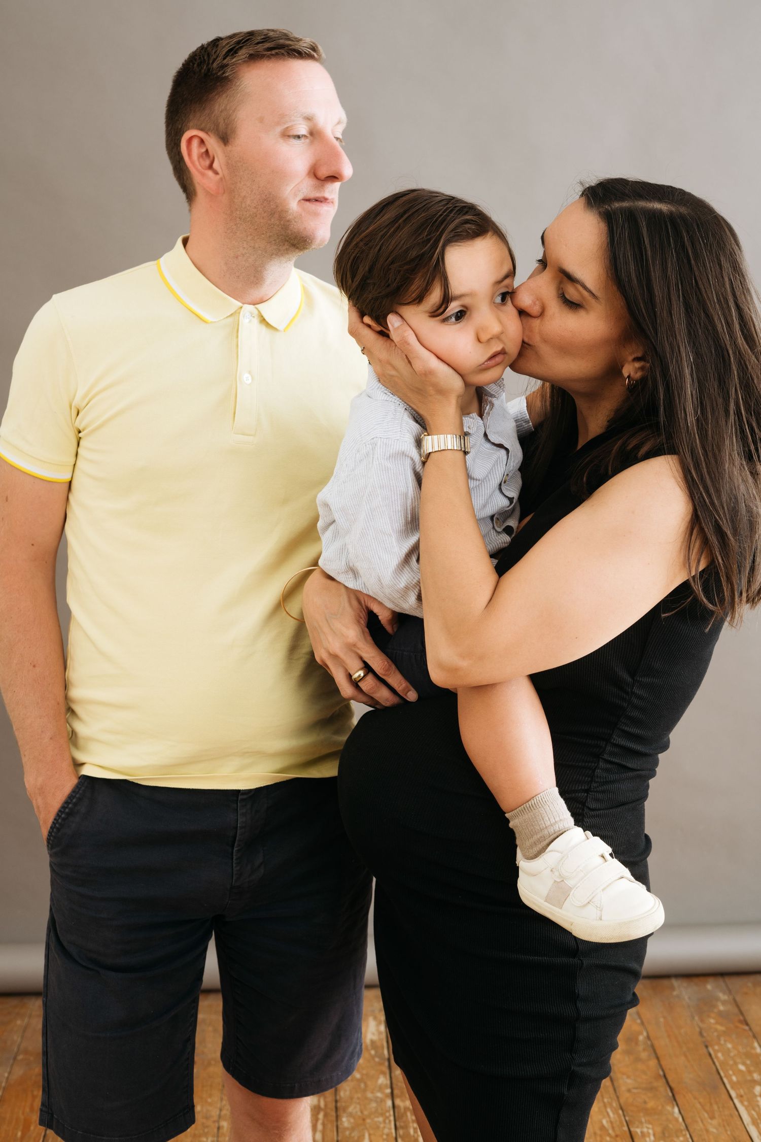 Family portrait of parents with young toddler son, mother kissing childs cheek in a studio setting.