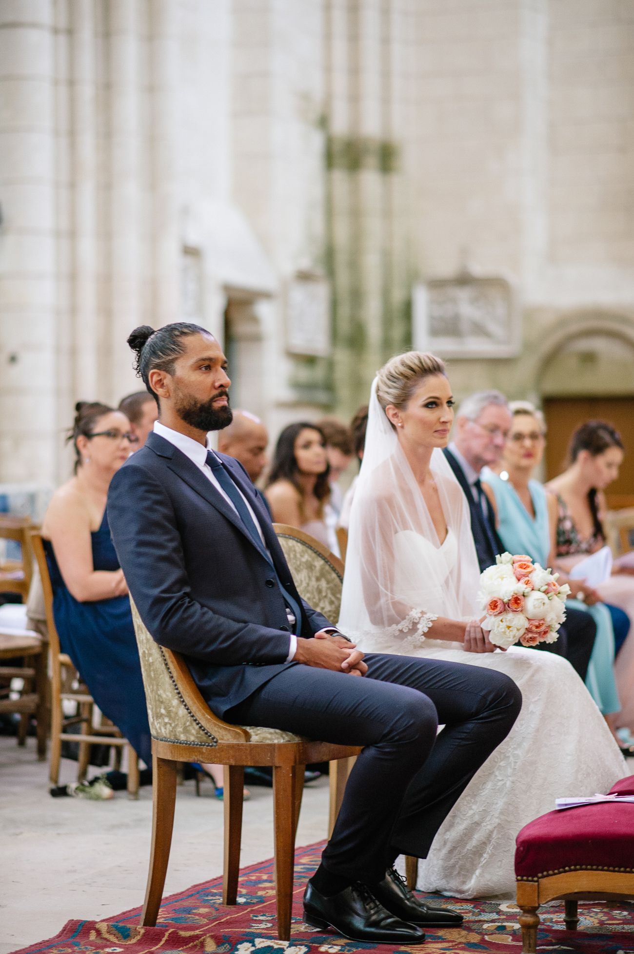 Bride and groom seated at church wedding ceremony, bride holding peach bouquet wearing veil, groom in navy suit.