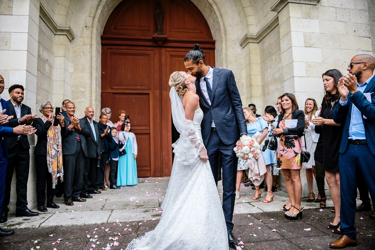 Newlywed couple sharing a kiss outside a church while wedding guests celebrate with confetti.