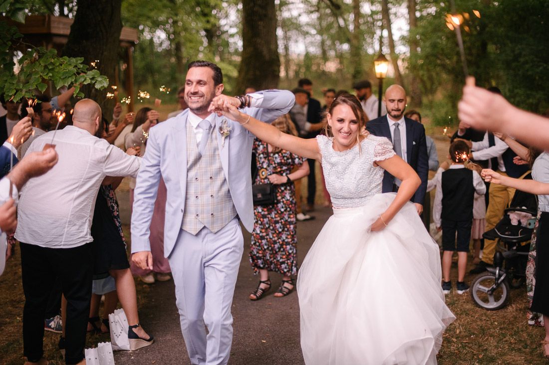 Joyful bride and groom walking through sparkler exit at outdoor wedding reception surrounded by cheering guests.