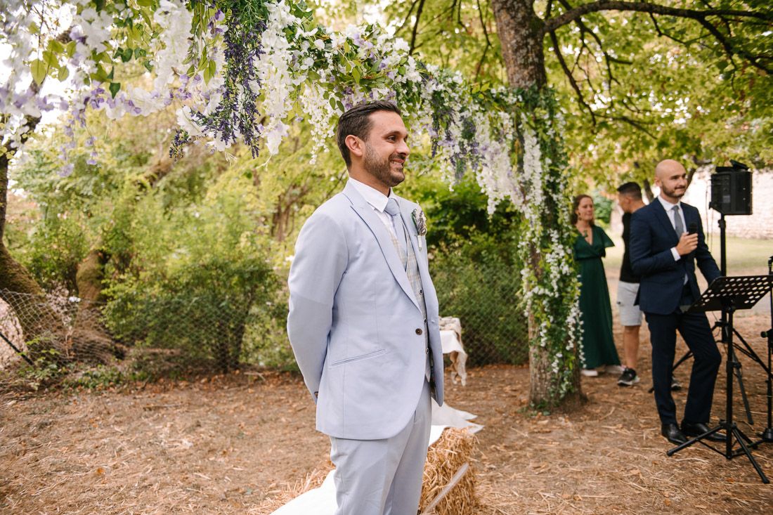 Smiling groom in light gray suit standing outdoors at a garden wedding ceremony with lush greenery.