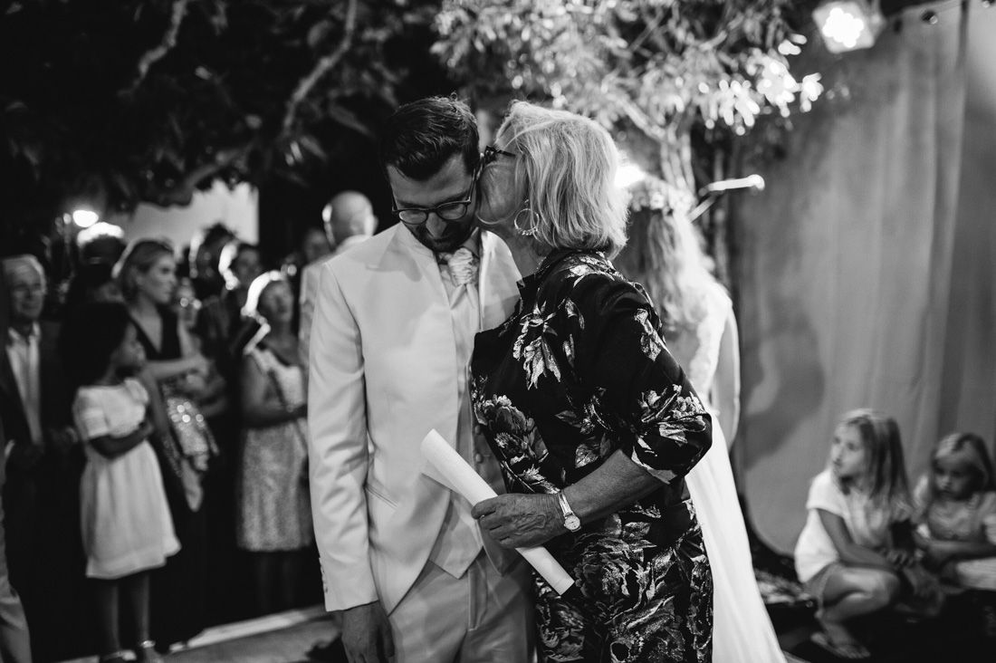 Black and white photo of a groom in white suit receiving a kiss on the cheek at an outdoor wedding reception.