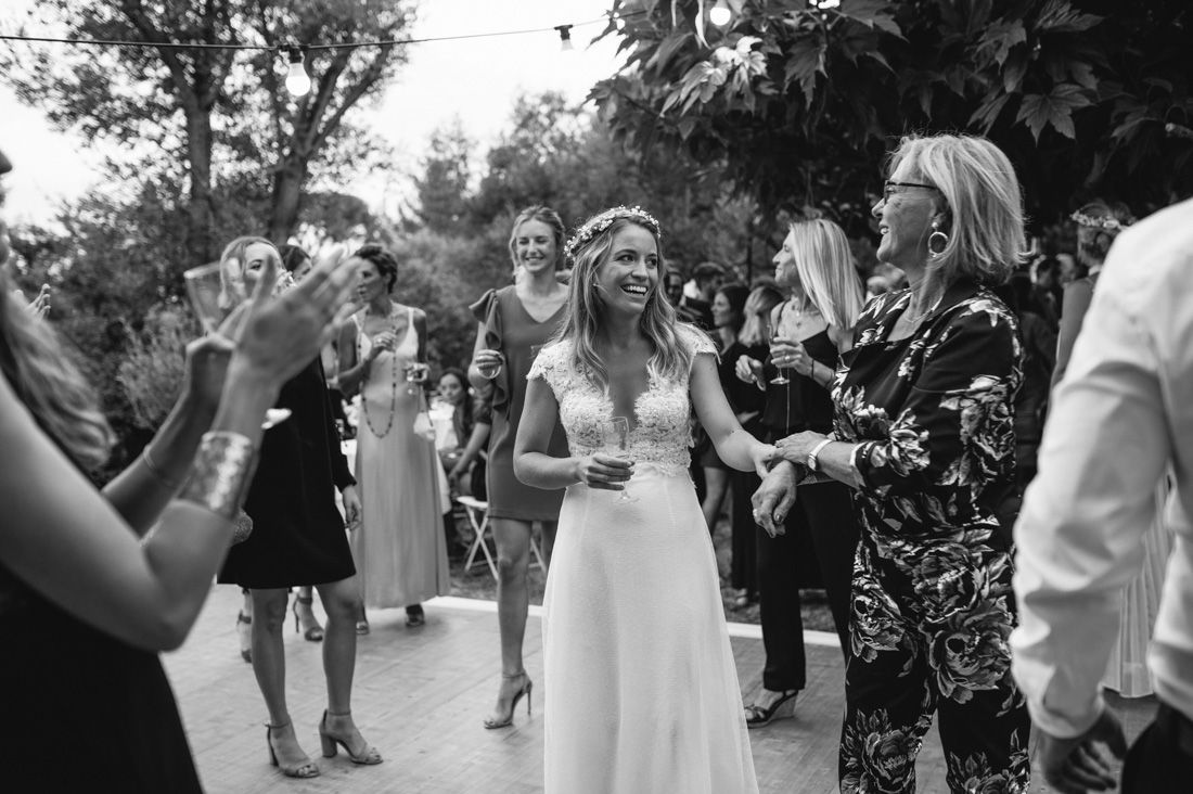 Bride in white gown with floral crown laughing and dancing with guests at outdoor wedding reception.