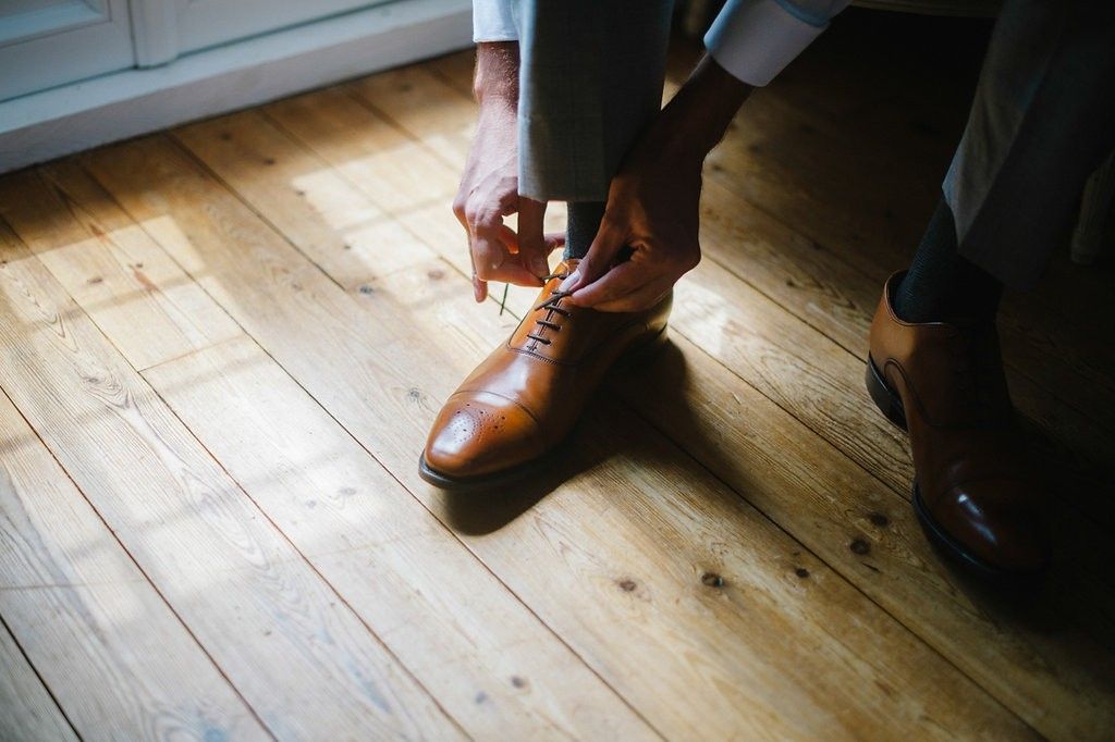 Groom tying brown leather oxford shoes on rustic hardwood floor before wedding.