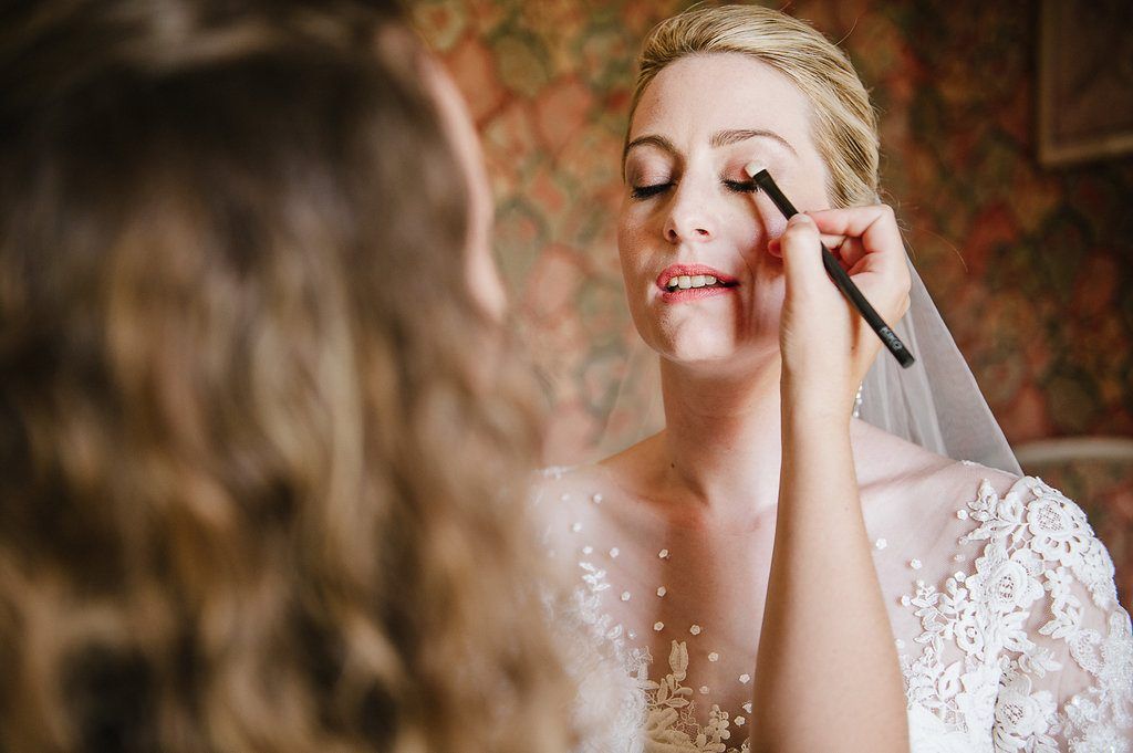 Makeup artist applying eye makeup to smiling bride wearing lace wedding dress and veil.