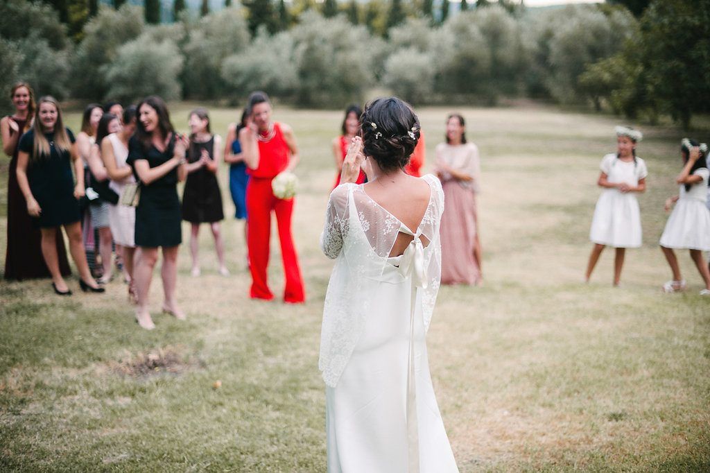 Bride in elegant backless lace wedding dress prepares to toss bouquet to waiting wedding guests outdoors.