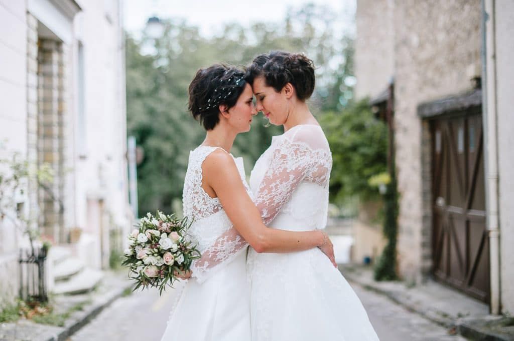 Two brides in white lace wedding dresses hold a bouquet and touch foreheads tenderly on a charming village street.