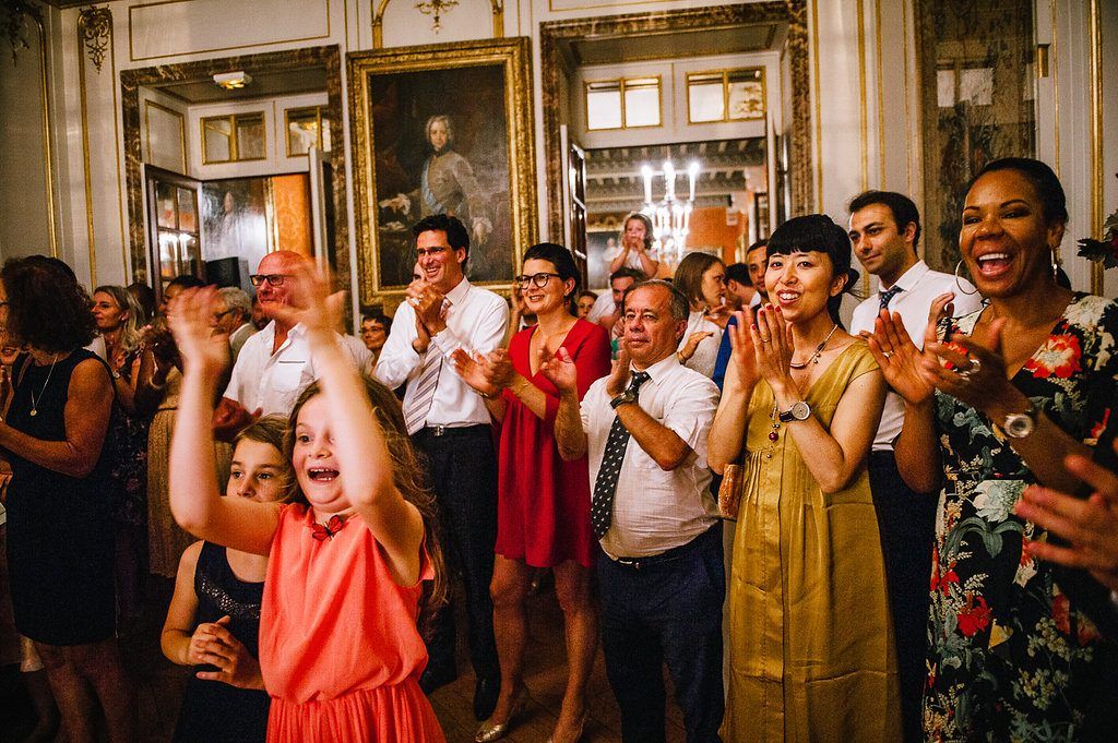 Joyful wedding guests clapping and celebrating during lively chateau reception with ornate gold decor.