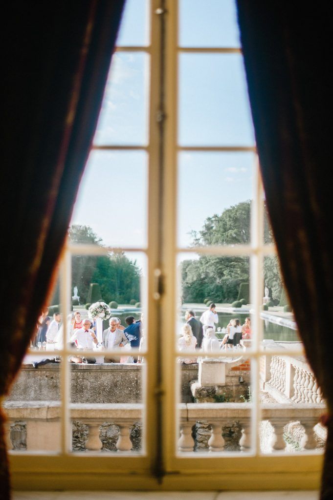 Wedding reception guests viewed through elegant French chateau windows with ornate stone balustrade.