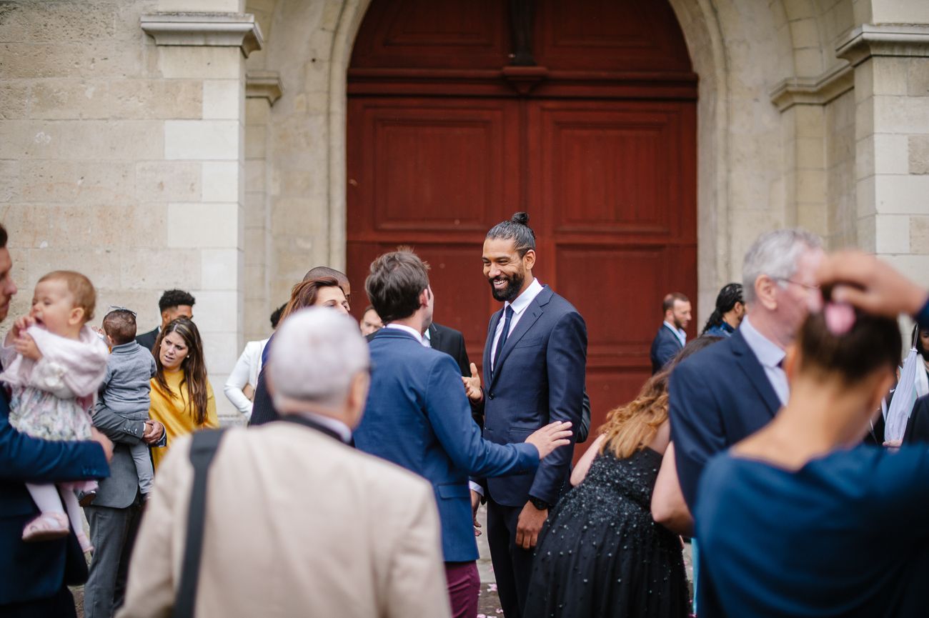 Groom in navy suit greeting wedding guests outside historic church entrance.