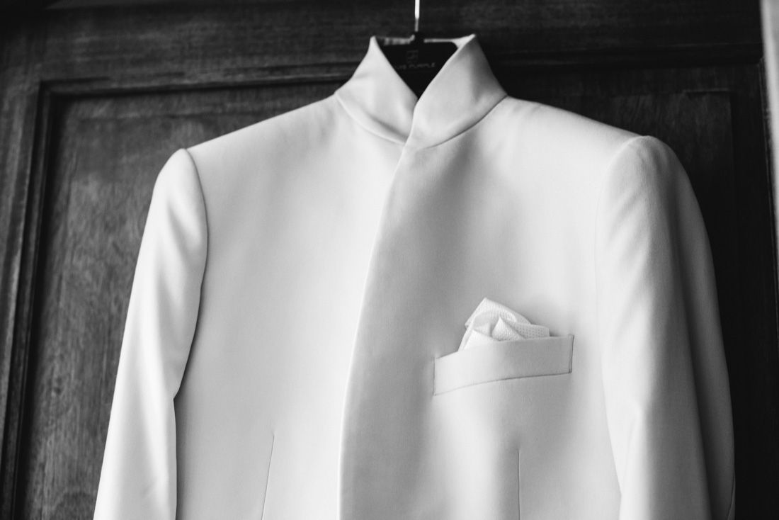 White mandarin collar suit jacket with pocket square hanging on wooden door in black and white photo.