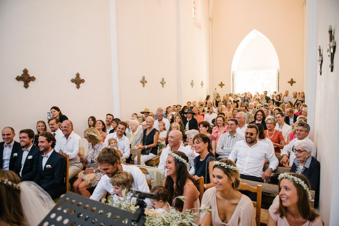 Guests seated in a white church during a wedding ceremony, with bridesmaids in blush dresses and floral crowns in the foreground.