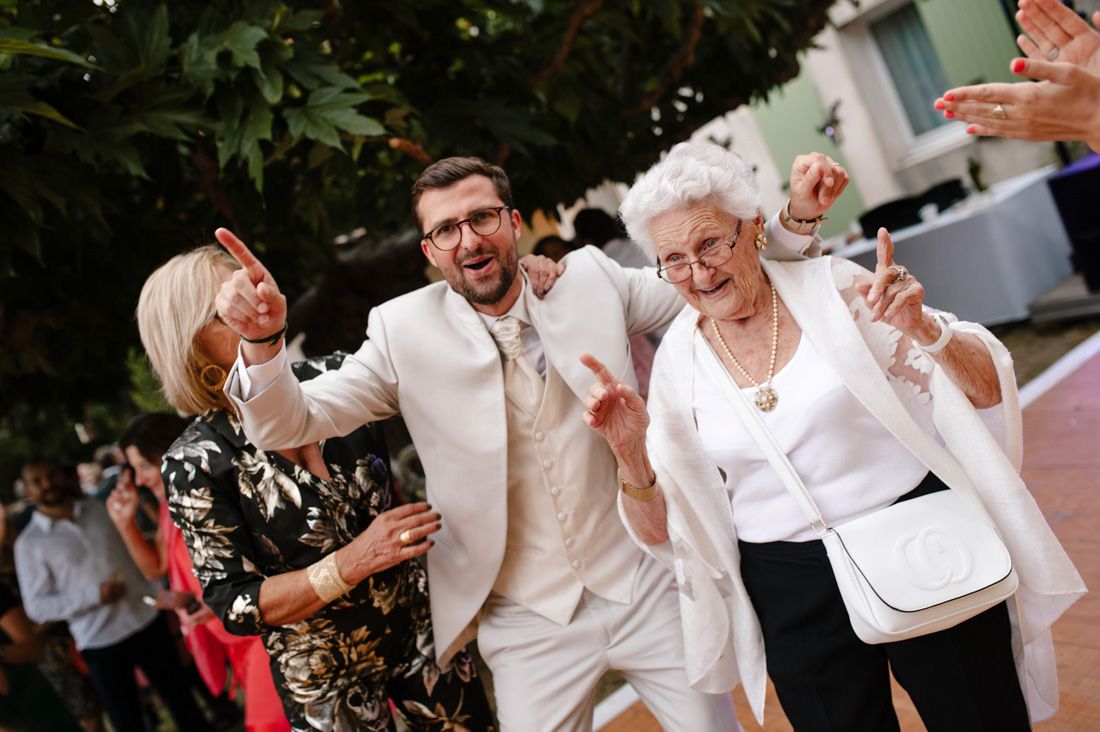 Groom in white suit dancing joyfully with elderly grandmother and guests at wedding reception.