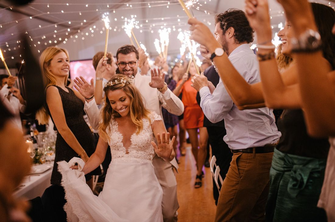 Bride walks through sparkler tunnel surrounded by cheering wedding guests during reception celebration.
