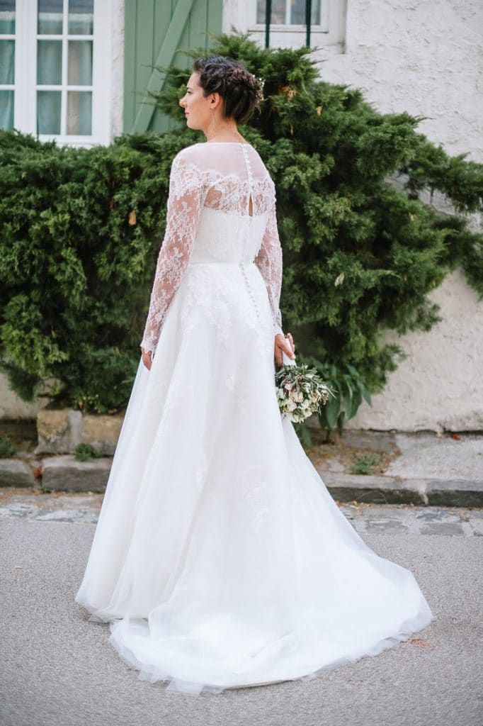 A bride in a long-sleeve lace wedding gown with a flowing skirt holds a bouquet while standing near green hedges.