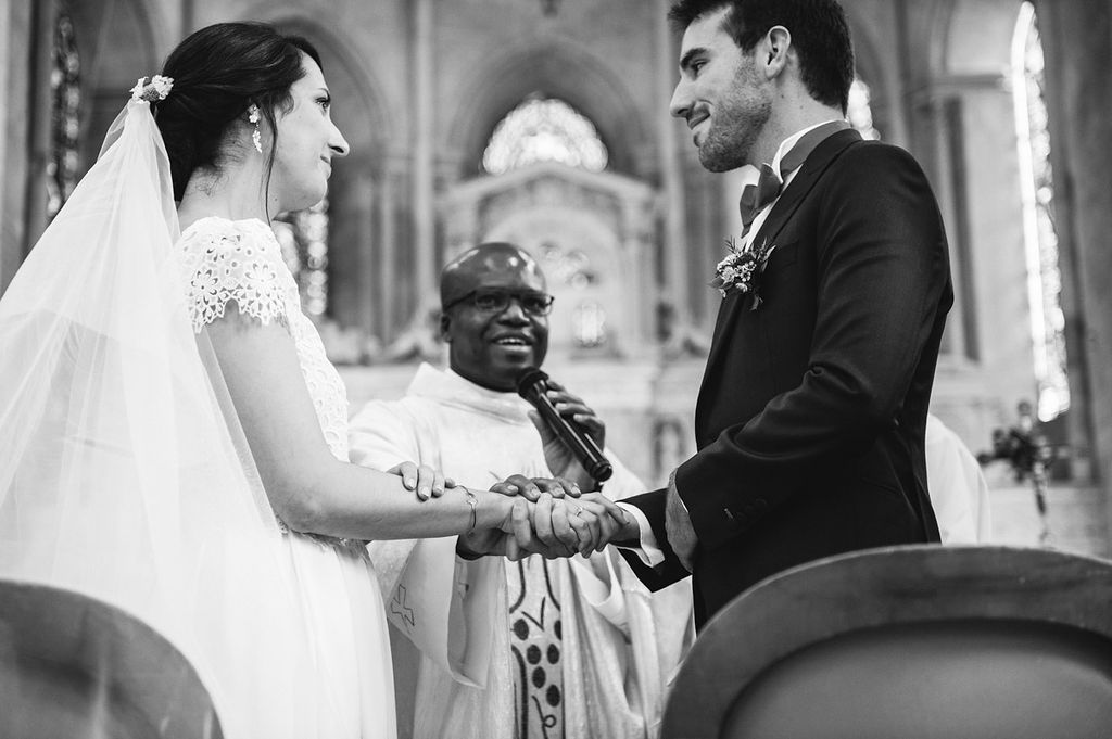 Black and white photo of bride and groom holding hands with officiant during church wedding ceremony.