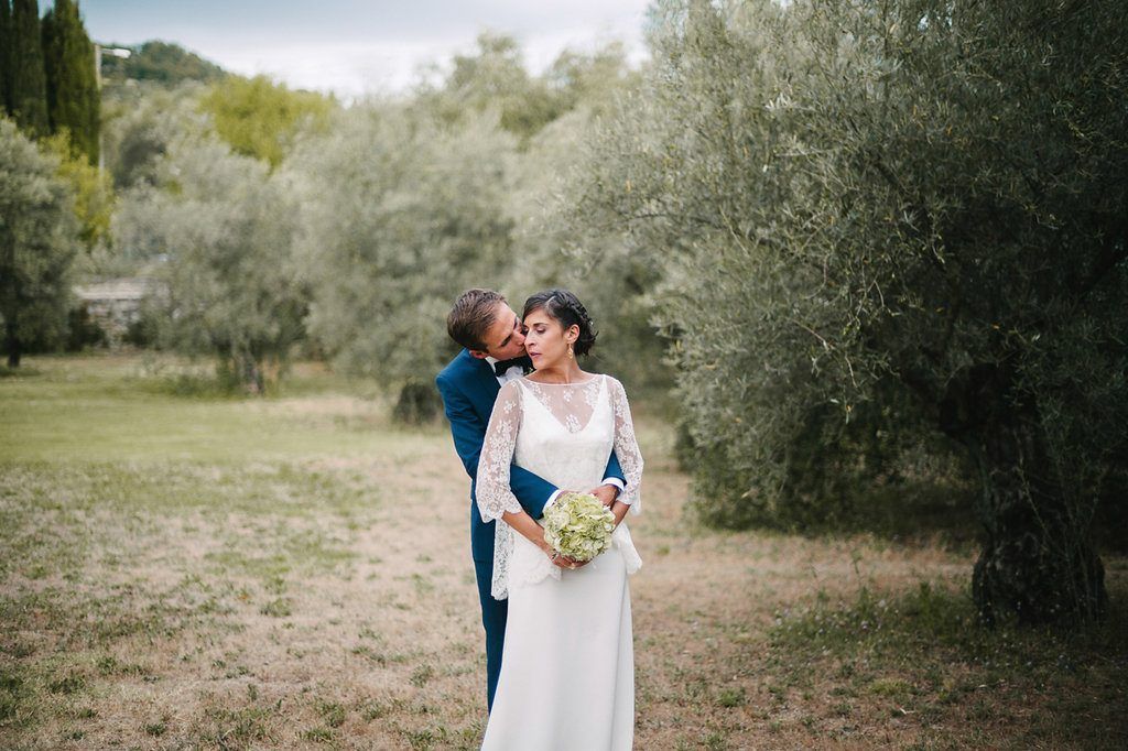 Groom in navy suit kisses bride on cheek as she smiles holding bouquet among olive trees.