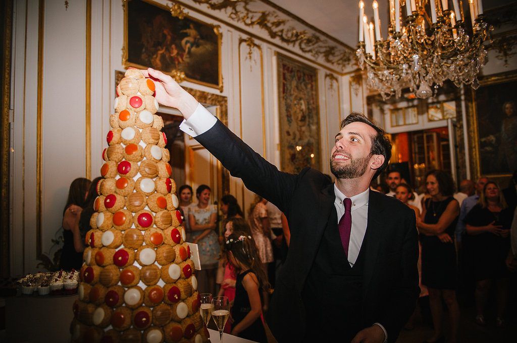 Groom in black suit reaches to top of elegant croquembouche wedding cake tower at ornate French chateau reception.