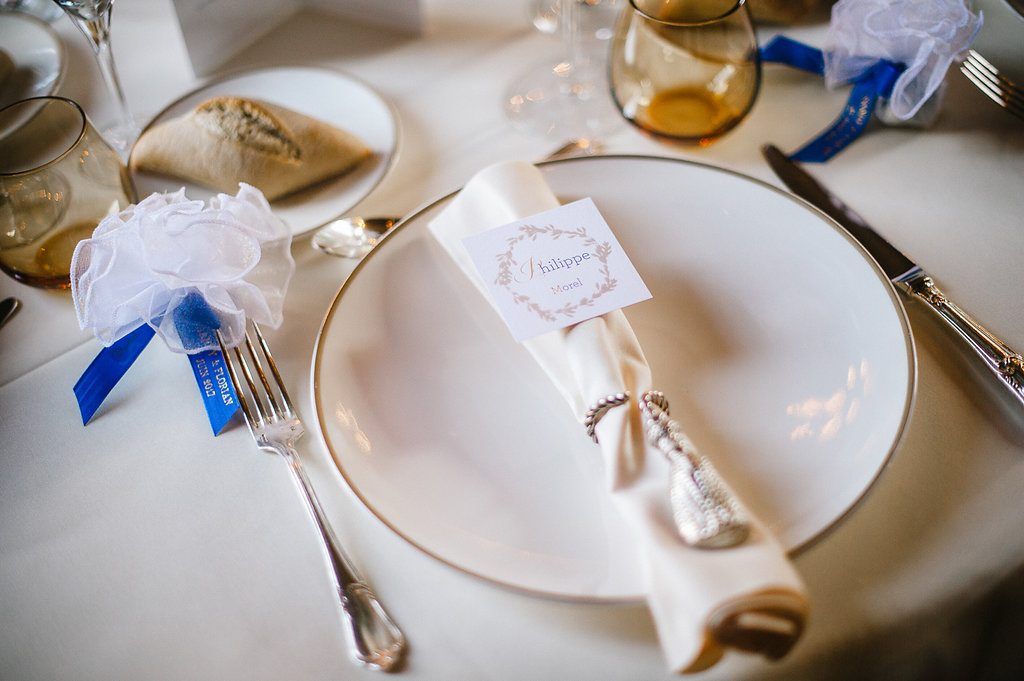 Elegant wedding place setting with ivory napkin, name card, blue ribbon favor, and gold-rimmed plate.