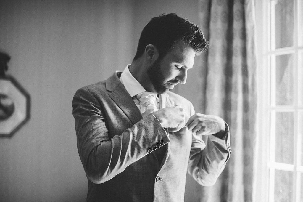 Black and white portrait of bearded groom in gray suit adjusting cufflinks by window.