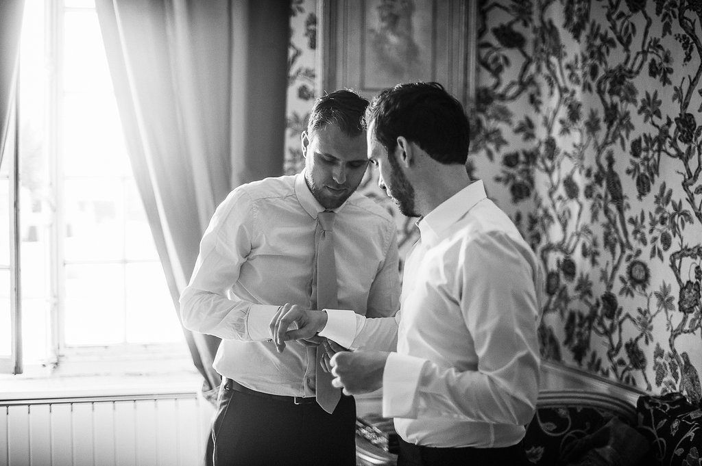 Two groomsmen in white shirts helping each other with cufflinks by window in ornate room.