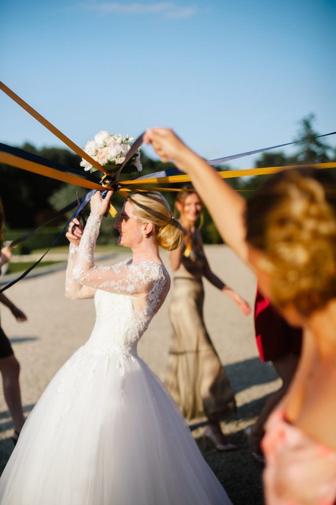 Bride in lace ballgown tosses ribbon-tied bouquet surrounded by bridesmaids in golden evening light.