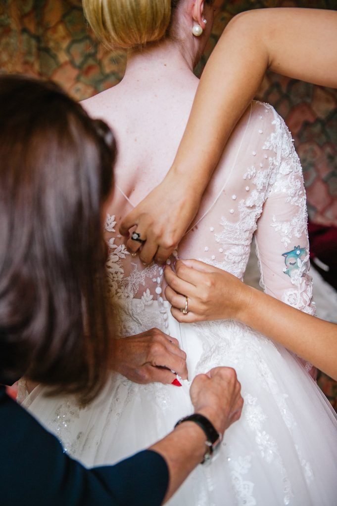 Two women helping bride fasten lace wedding dress with floral embroidery details from behind.