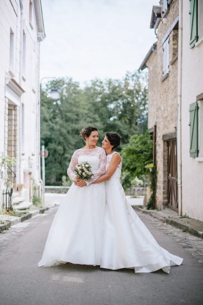 Two brides in white ball gowns embrace on a quiet village street holding a bouquet of white and pink flowers.