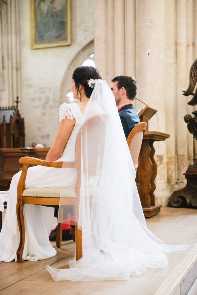 Bride in lace gown with long veil sits beside groom during their church wedding ceremony.