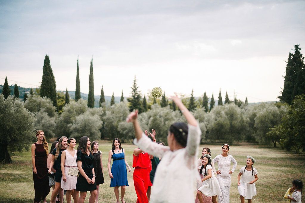 Bride in white lace gown tosses bouquet to group of guests on green lawn with cypress trees.