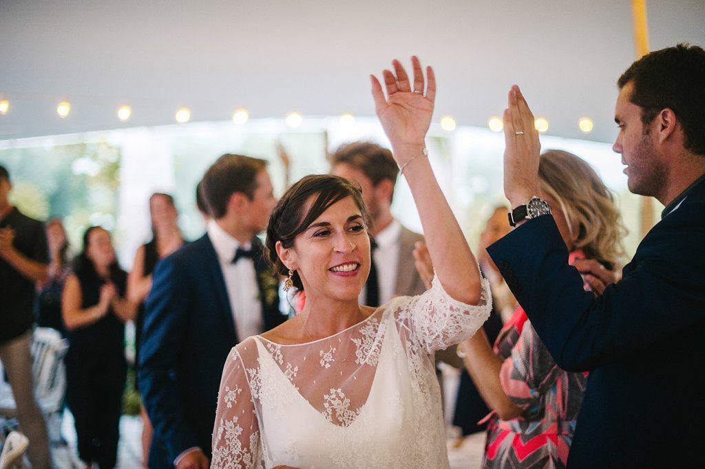 Smiling bride in lace wedding dress raising hand while dancing at wedding reception.